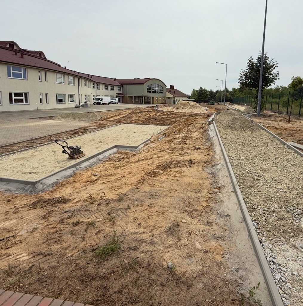 Przygotowanie terenu pod budowę: ubijarka w piasku, obrzeża chodnika z kamieniami, budynek w tle. Etap prac ziemnych z użyciem sprzętu budowlanego.