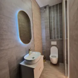 Nowoczesna łazienka w szarościach z podświetlanym lustrem, umywalką nablatową i toaletą. Minimalistyczny design, płytki imitujące kamień.