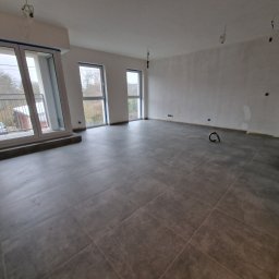 R-Art Wykończenia Wnętrz - Szare płytki podłogowe w nowym mieszkaniu z dużymi oknami balkonowymi i widokiem na zewnątrz. Ściany przygotowane do malowania. Widoczne niedokończone elementy instalacji.