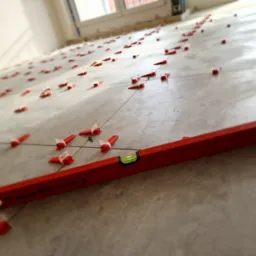 Ułożone płytki podłogowe z systemem poziomowania, widoczna poziomica. Jasne wnętrze, neutralna kolorystyka płytek.