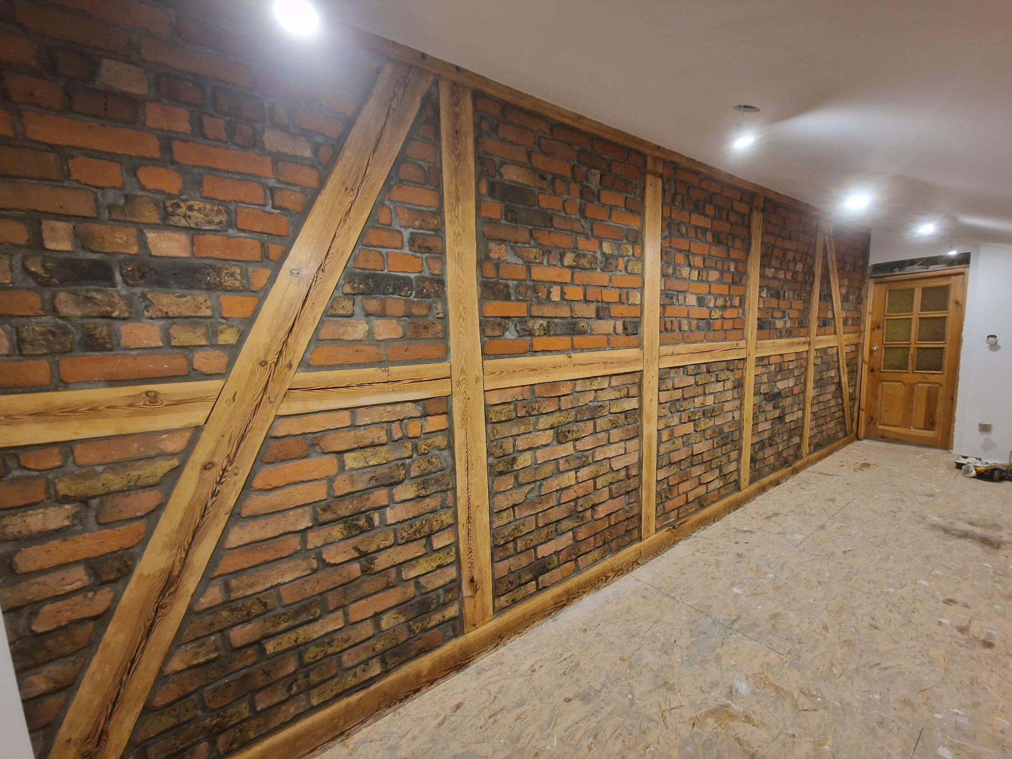 Ściana z cegły klinkierowej w rustykalnym stylu z drewnianą konstrukcją szkieletową. Widoczna podłoga z płyt OSB i drewniane drzwi w tle.
