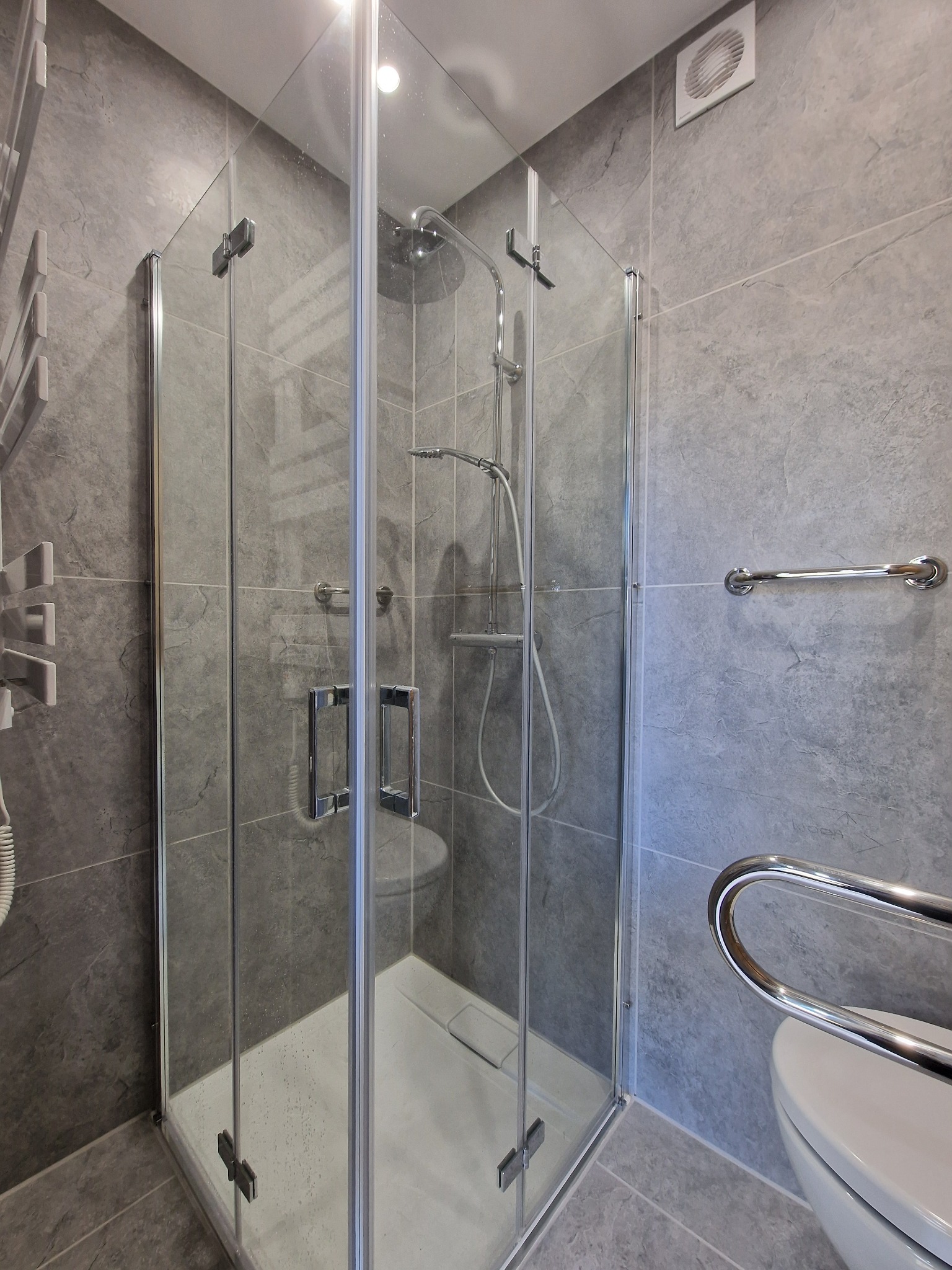 Szara łazienka z kabiną prysznicową typu walk-in i chromowanymi uchwytami dla niepełnosprawnych, widok na płytki imitujące kamień.