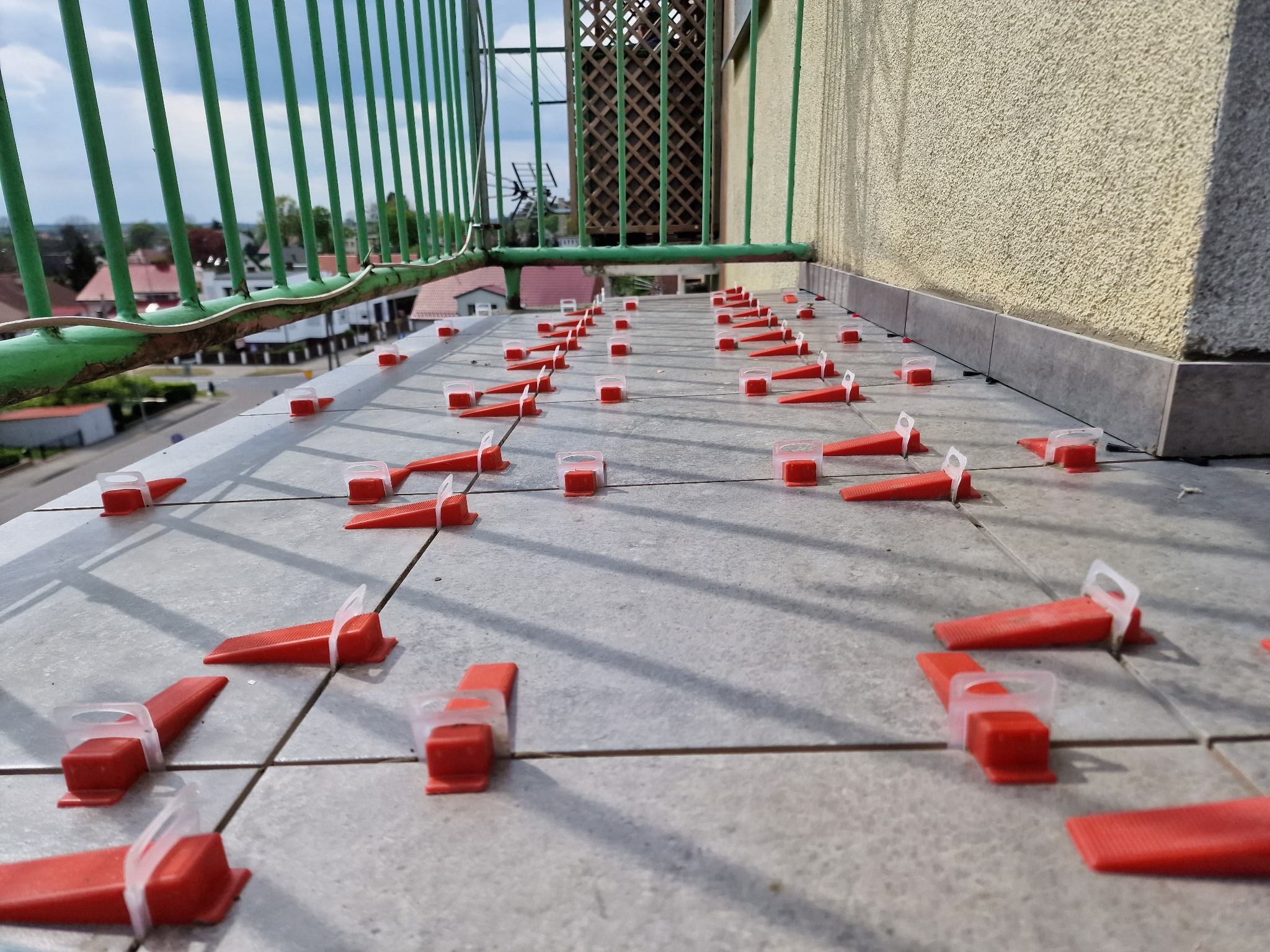 Kafelki na balkonie w trakcie układania, widoczne czerwone kliny i klipsy poziomujące. W tle zielona barierka balkonu i zabudowa miejska.