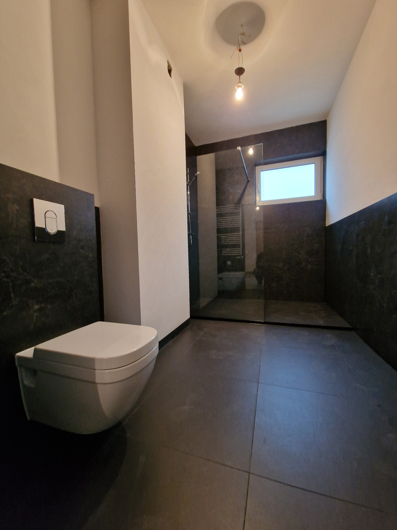 Nowoczesna łazienka po remoncie: czarne płytki, podwieszana toaleta z chromowanym przyciskiem, przeszklona kabina prysznicowa i sufit z wystającym okablowaniem.