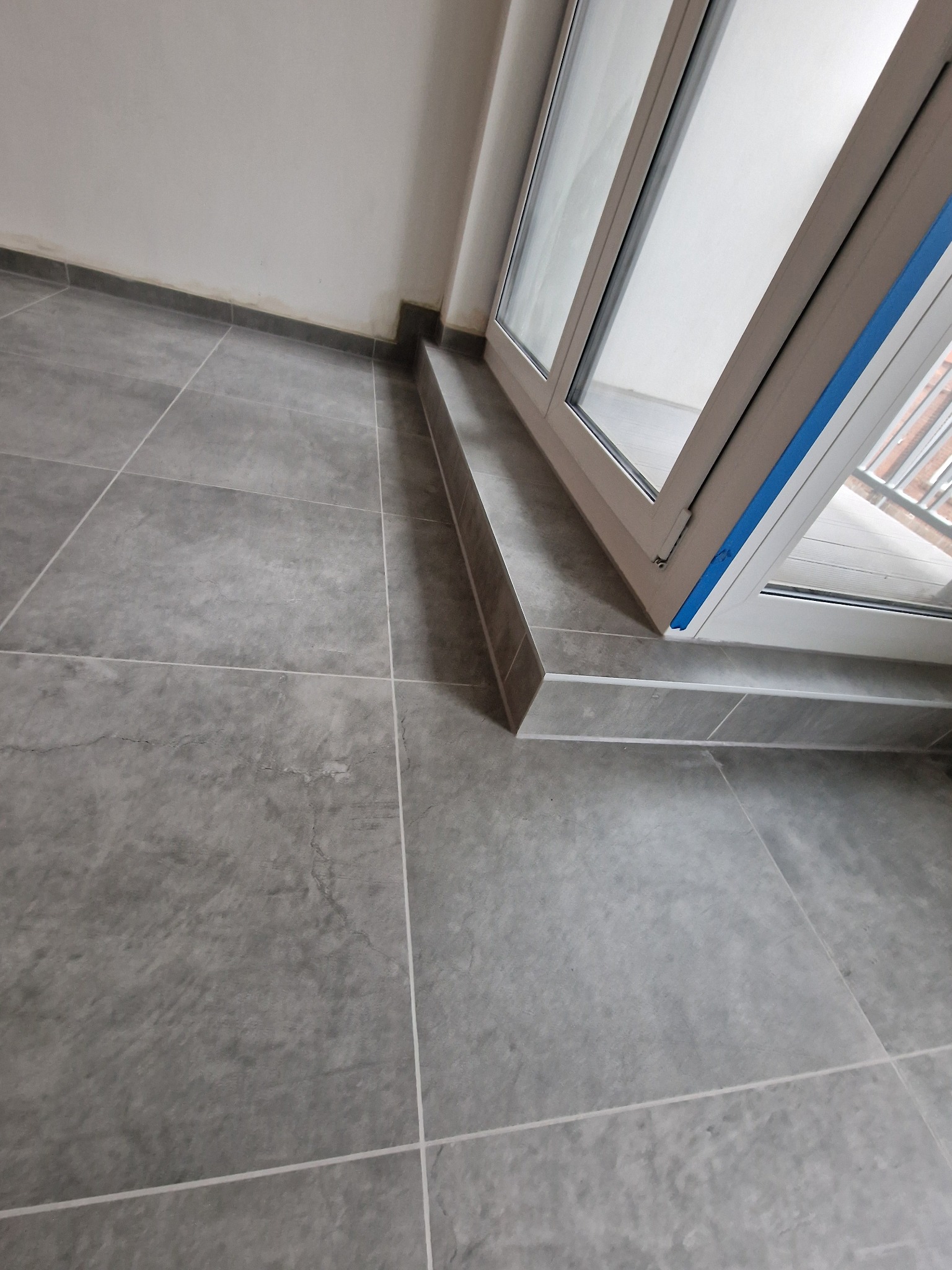 Szare płytki podłogowe ułożone w kwadraty, widoczne przy oknie balkonowym z niebieską taśmą zabezpieczającą. Minimalistyczne wykończenie.