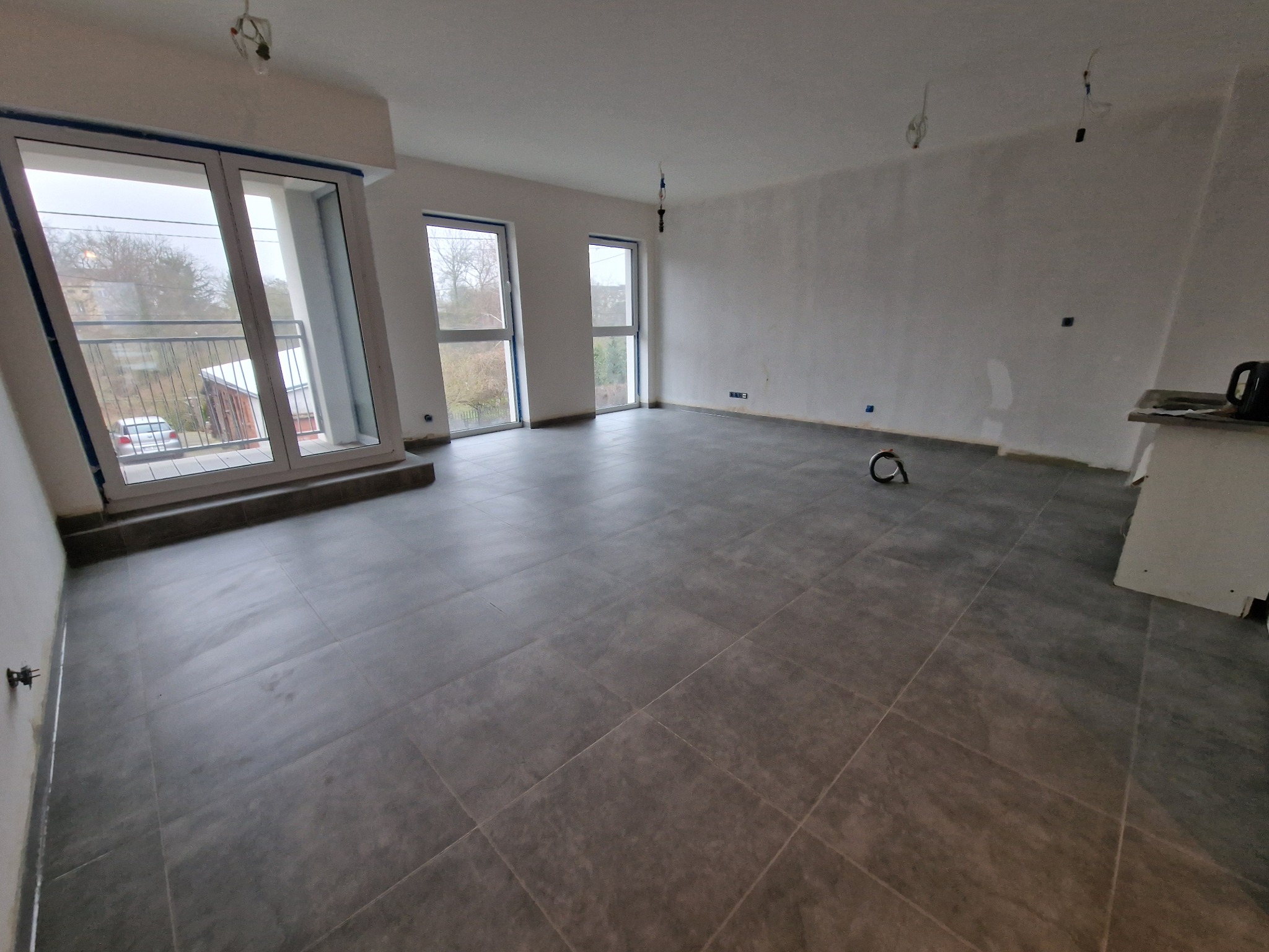 Szare płytki podłogowe w nowym mieszkaniu z dużymi oknami balkonowymi i widokiem na zewnątrz. Ściany przygotowane do malowania. Widoczne niedokończone elementy instalacji.