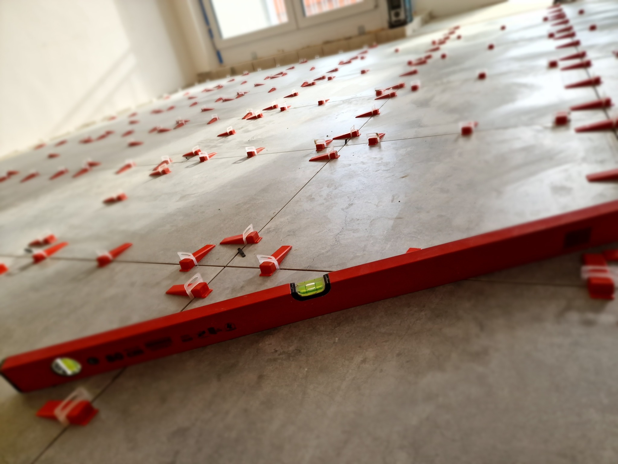 Ułożone płytki podłogowe z systemem poziomowania, widoczna poziomica. Jasne wnętrze, neutralna kolorystyka płytek.