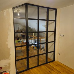 Ases sp zoo - Loftowa ścianka działowa z czarnymi szprosami i przezroczystym szkłem, oddzielająca przestrzeń w domu. Widoczny drewniany parkiet i fragment ściany w trakcie wykończenia.