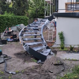 Ases sp zoo - Montaż czarnych, spiralnych schodów metalowych na zewnątrz budynku, zabezpieczonych folią. Widoczne rusztowanie i narzędzia.