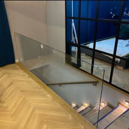 Ases sp zoo - Nowoczesna balustrada szklana na schodach z oświetleniem LED, widok z góry. Parkiet w jodełkę i czarna rama okienna w tle.