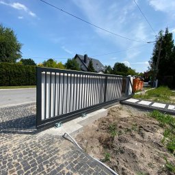 Ases sp zoo - Nowoczesna brama przesuwna z czarnymi, pionowymi profilami na tle białego muru, widoczna z poziomu brukowanej ścieżki w słoneczny dzień.