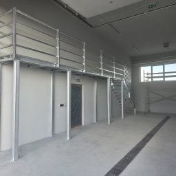 Ases sp zoo - Wnętrze hali z metalową konstrukcją antresoli, schodami i balustradą. Widoczne słupy nośne, barierki zabezpieczające i kratka ściekowa w posadzce. Surowe wykończenie.