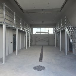 Ases sp zoo - Wnętrze hali z metalową konstrukcją antresoli i schodami. Widoczne metalowe słupy, balustrady i schody z kratownicy. Surowy, industrialny charakter przestrzeni.