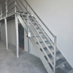 Ases sp zoo - Metalowe schody z ażurowymi stopniami i balustradą, prowadzące na antresolę. Widoczne mocowania śrubowe i surowy, industrialny charakter konstrukcji.