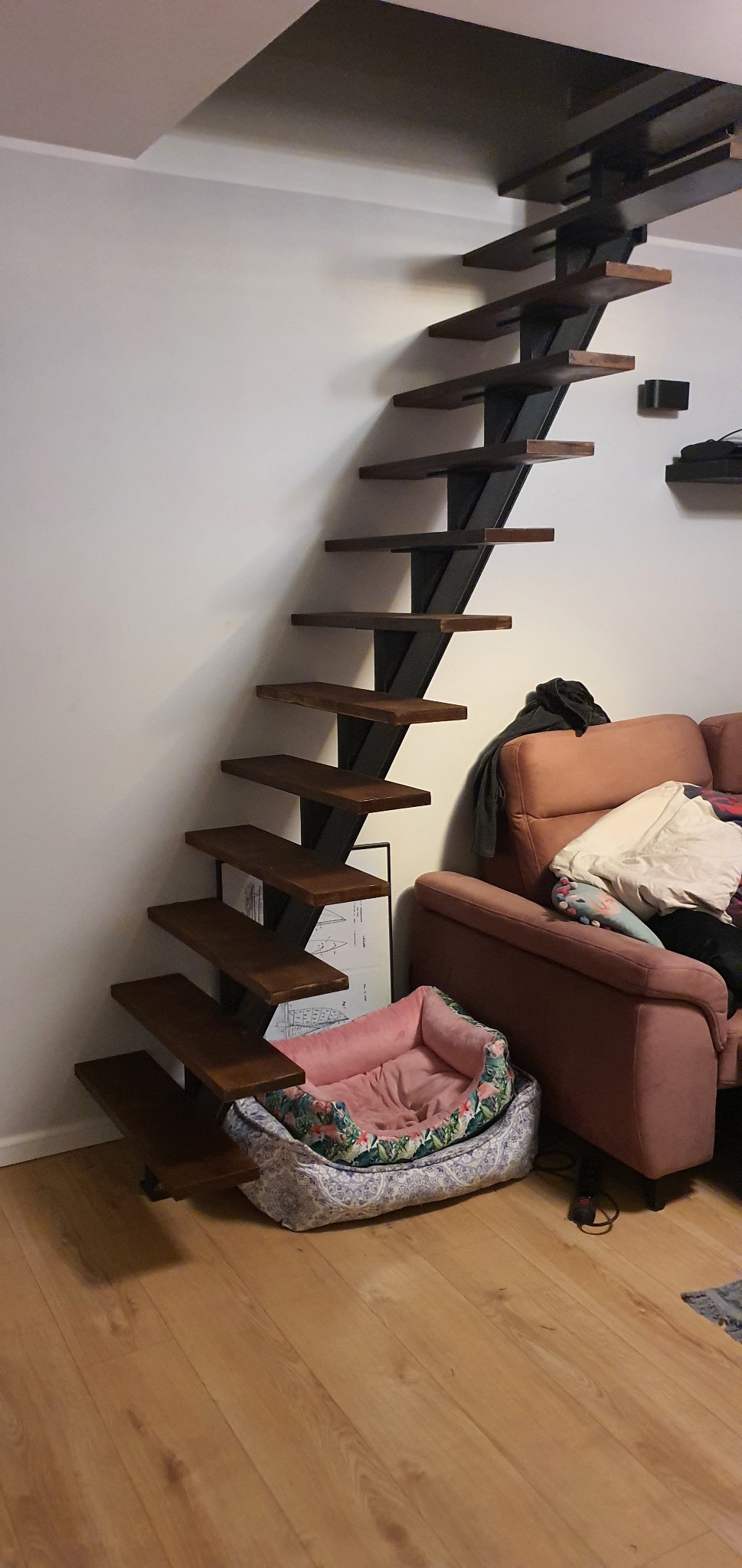 Drewniane schody wspornikowe z czarną metalową konstrukcją, zamontowane przy białej ścianie w pokoju z kanapą i legowiskiem dla zwierząt.