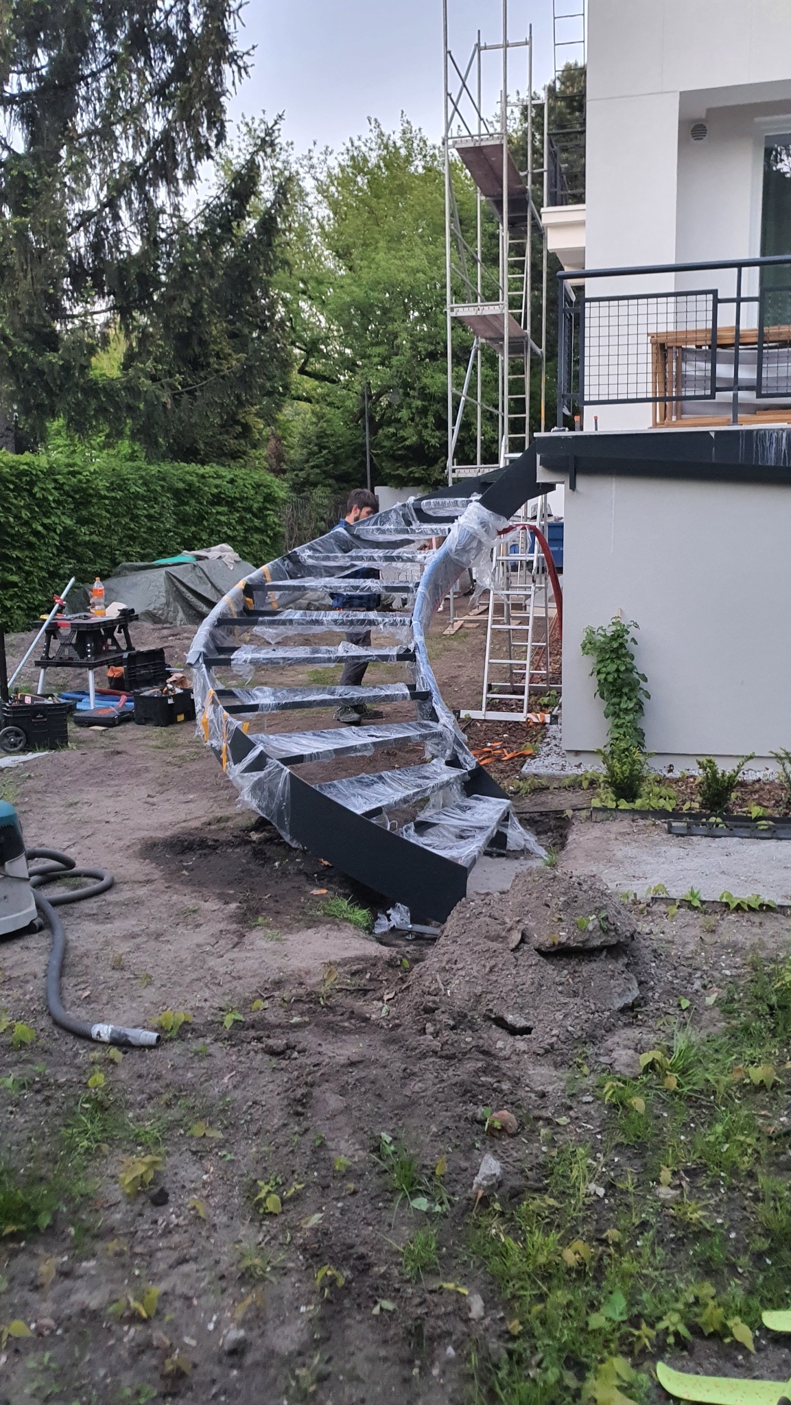Montaż czarnych, spiralnych schodów metalowych na zewnątrz budynku, zabezpieczonych folią. Widoczne rusztowanie i narzędzia.