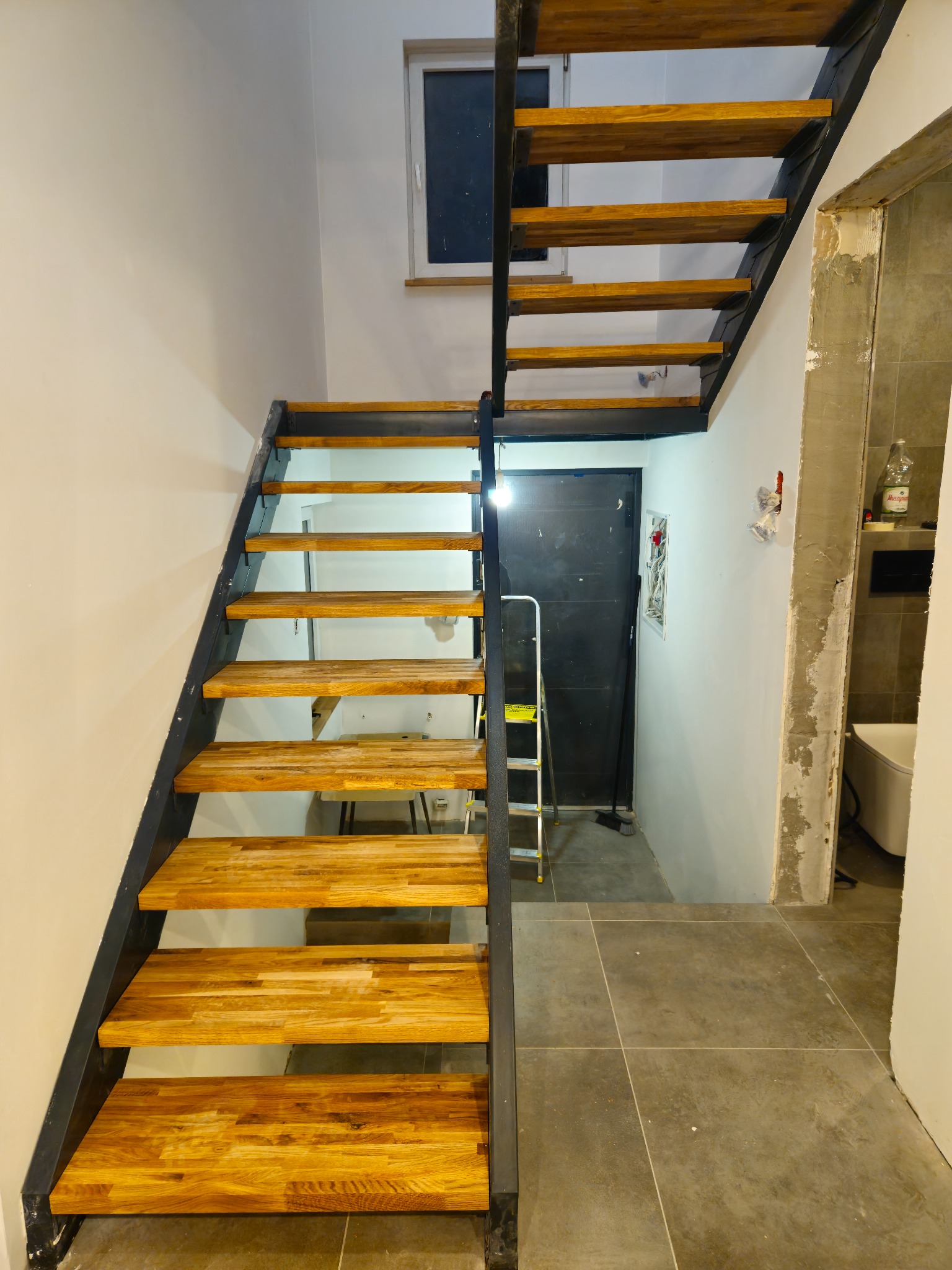 Metalowe schody z drewnianymi stopniami w jasnym wnętrzu. Widoczna drabina i wejście do łazienki w trakcie wykończenia. Minimalistyczny design, surowe wykończenie.