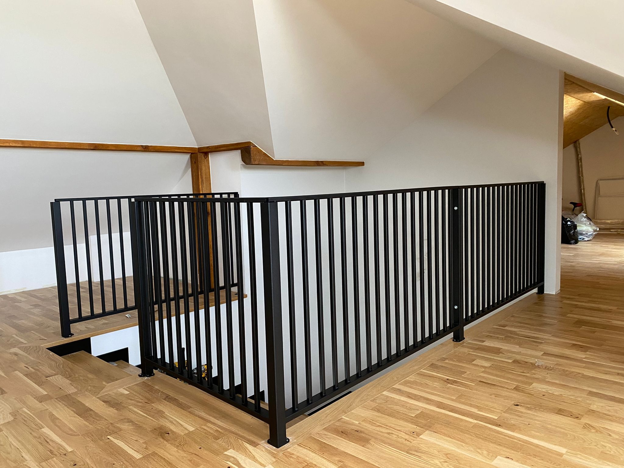 Czarna, metalowa balustrada schodowa z pionowymi szczebelkami na drewnianej podłodze. Widok z góry, proste linie, nowoczesny design, białe ściany, naturalne światło.