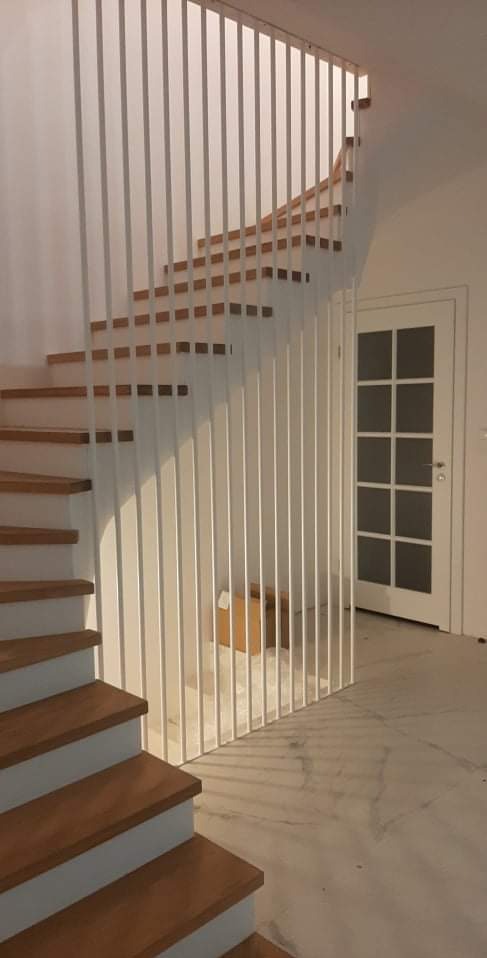 Nowoczesne schody drewniane z białą, minimalistyczną balustradą pionową w jasnym wnętrzu, widok z dołu na pierwsze stopnie i fragment drzwi.