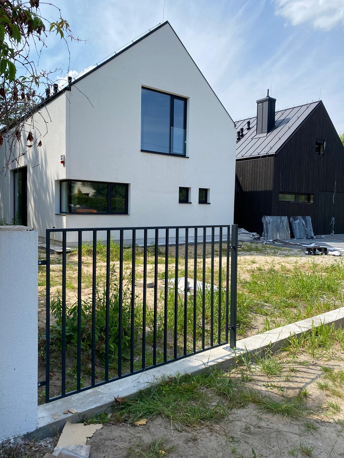 Nowoczesne ogrodzenie panelowe w kolorze czarnym, oddzielające posesję z dwoma domami o minimalistycznej architekturze. Widok na trawnik i fragmenty materiałów budowlanych.