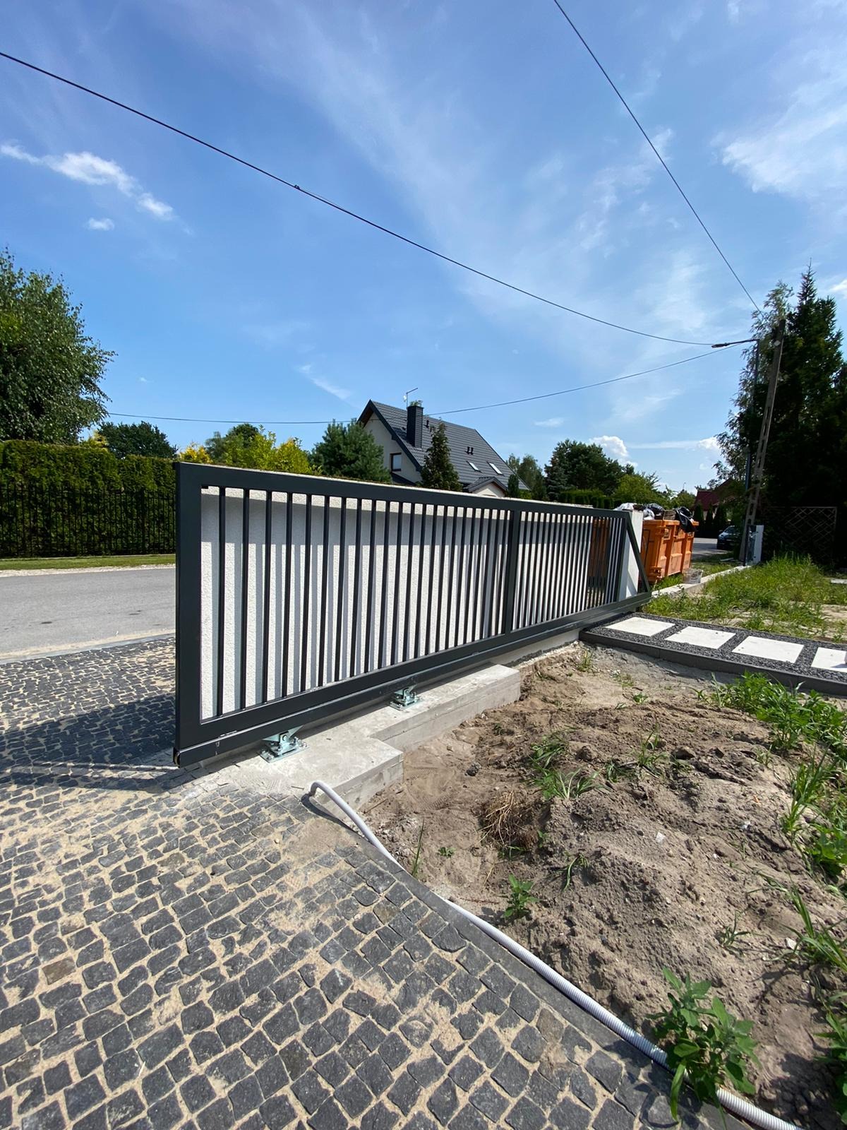Nowoczesna brama przesuwna z czarnymi, pionowymi profilami na tle białego muru, widoczna z poziomu brukowanej ścieżki w słoneczny dzień.