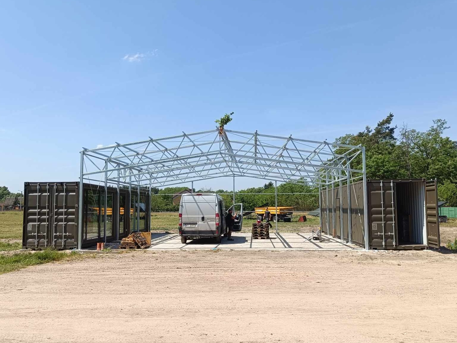 Konstrukcja metalowa z kontenerami po bokach, prawdopodobnie w trakcie budowy, na zewnątrz, w tle widoczny samochód dostawczy i drzewa.
