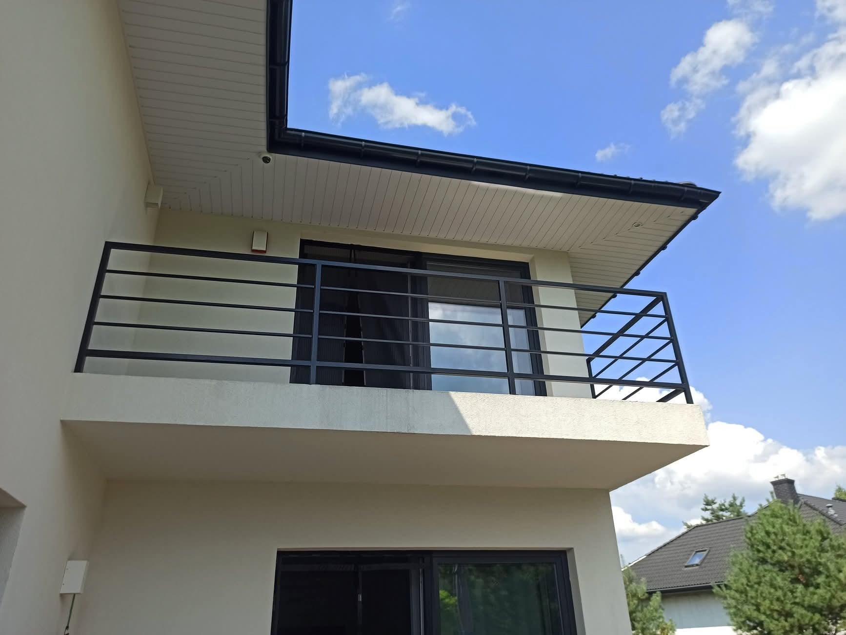 Elegancka, czarna balustrada balkonowa z poziomymi prętami, zamontowana na jasnym balkonie budynku z widokiem na błękitne niebo z chmurami. Minimalistyczny design.