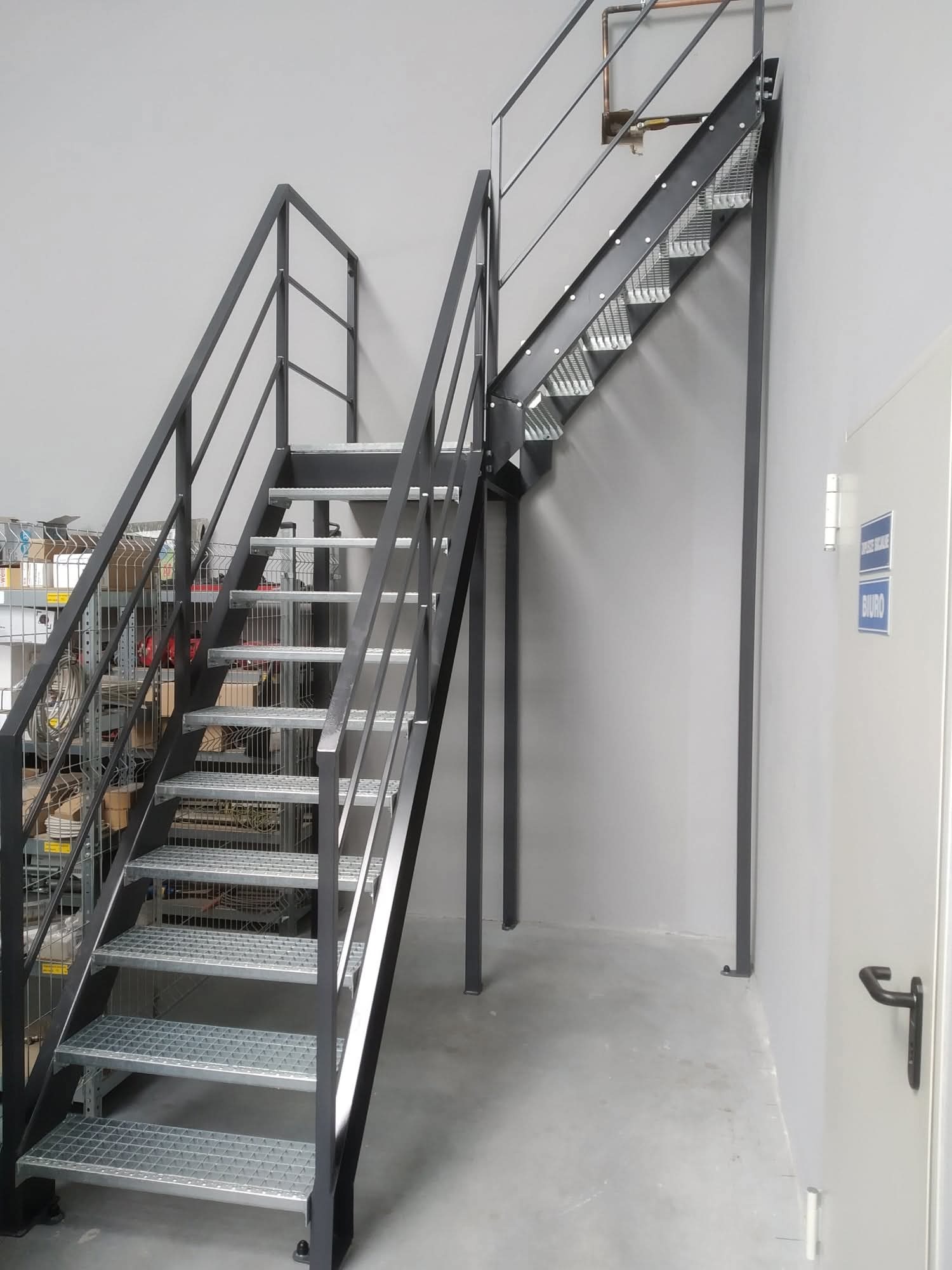 Metalowe schody z ażurowymi stopniami i czarną balustradą w szarym wnętrzu biurowym. Widoczne regały w tle i znak z napisem 'Biuro' na drzwiach.