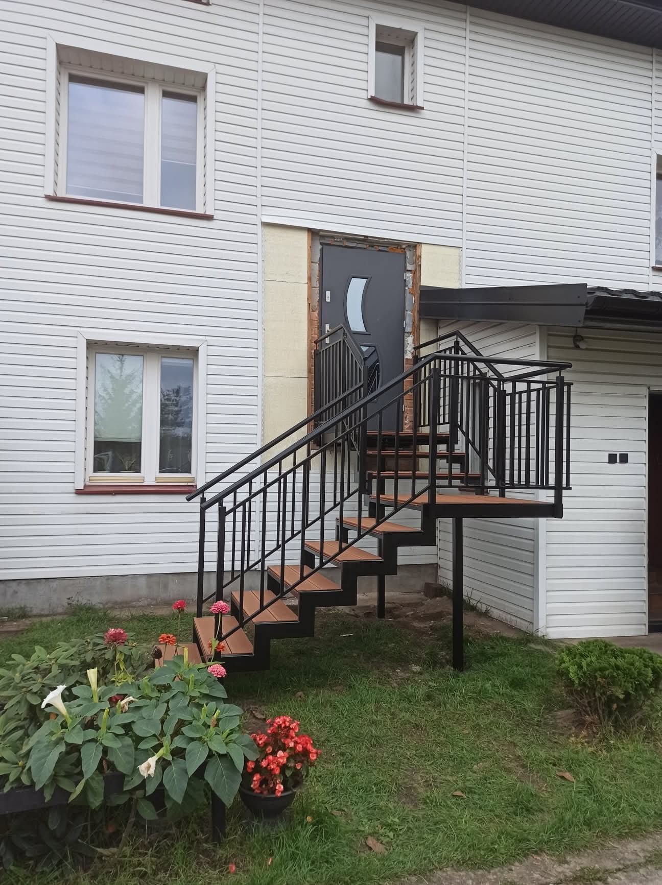 Zewnętrzne schody metalowe z czarną balustradą i drewnianymi stopniami prowadzą do drzwi domu z sidingiem. Na pierwszym planie kwiaty.
