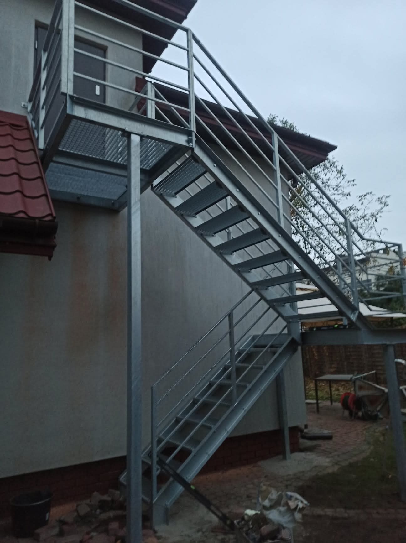 Zewnętrzne schody metalowe z balustradą, prowadzące do drzwi na piętrze budynku. Konstrukcja ocynkowana, z ażurowymi stopniami. Widok z dołu, na tle elewacji i fragmentu dachu.