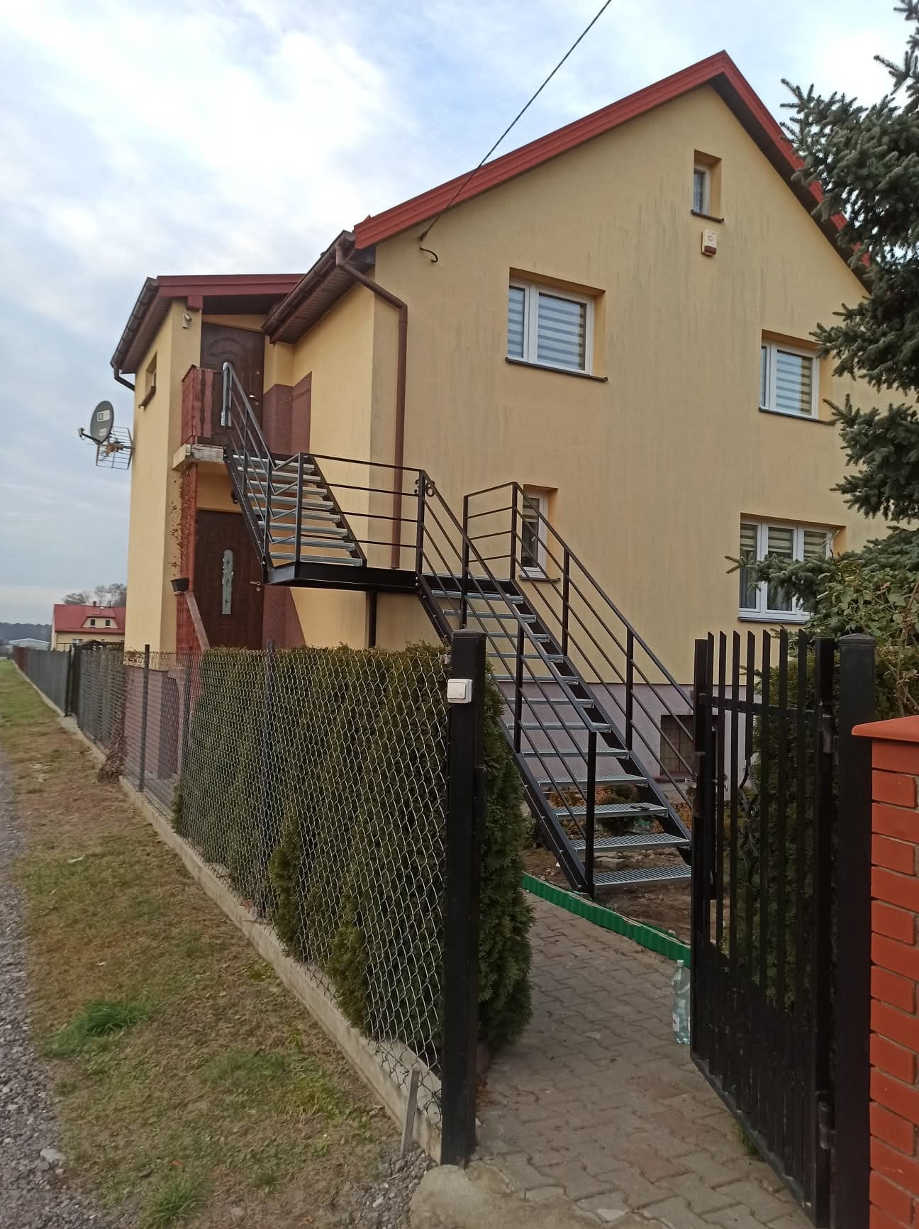 Żółty dom z zewnętrznymi schodami metalowymi prowadzącymi na balkon. Czarna brama i ogrodzenie z siatki otaczają posesję. Widok na elewację i elementy metalowe.