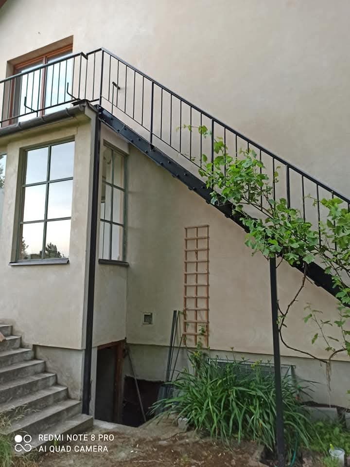 Zewnętrzne schody metalowe z czarną balustradą prowadzące na balkon, z roślinami pnącymi się po balustradzie. Widok na jasną elewację budynku i okna.