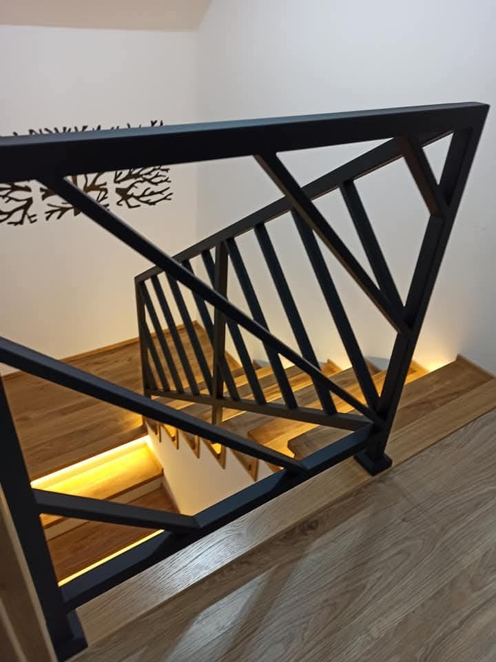 Nowoczesna, czarna balustrada schodowa z geometrycznym wzorem i drewnianymi schodami z podświetleniem LED, widziana z góry pod kątem. Minimalistyczny design.