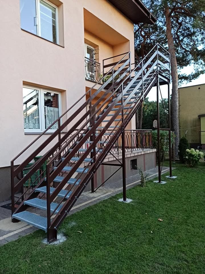 Zewnętrzne schody metalowe z brązową balustradą prowadzące na balkon, widoczne z poziomu trawnika, z elementami kotwienia w betonie.