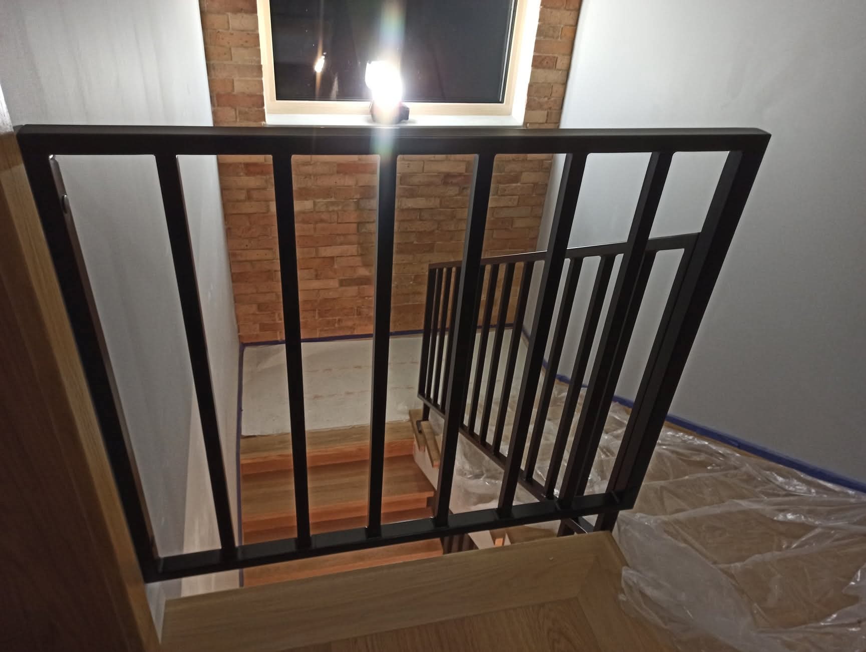 Nowoczesna, czarna balustrada metalowa przy schodach wewnątrz budynku, Góra Kalwaria. Widoczne drewniane stopnie i ceglana ściana w tle. Ochrona podłogi folią malarską.