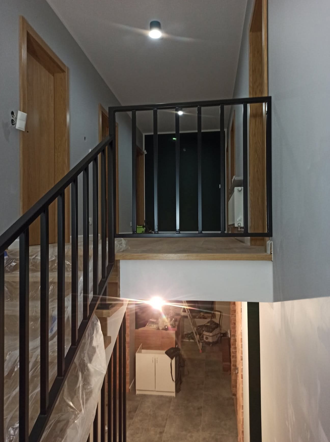 Czarne, metalowe schody z balustradą wewnątrz domu. Schody zabezpieczone folią, widoczne drzwi i fragment pomieszczenia poniżej schodów. Minimalistyczny design.