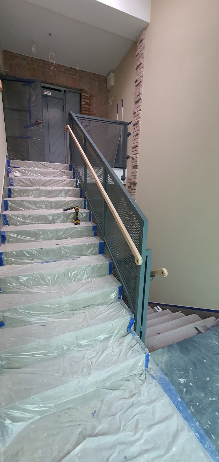 Metalowe schody z ażurową balustradą w trakcie montażu. Schody zabezpieczone folią, widoczny wkrętak akumulatorowy. Surowa ściana z cegły w tle.