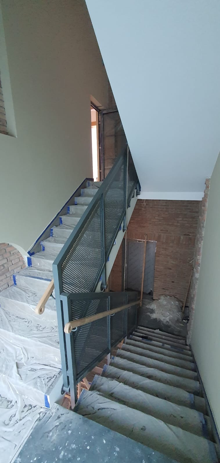 Metalowe schody z ażurową balustradą i drewnianym poręczą w trakcie montażu. Schody zabezpieczone folią malarską. Widok z góry na dół.