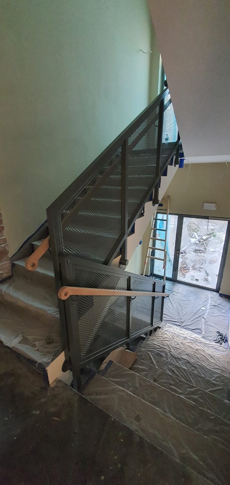 Nowoczesne schody z metalową, ażurową balustradą i drewnianym pochwytem, widoczne zabezpieczenia na schodach, drabina w tle, wnętrze w trakcie wykończenia.