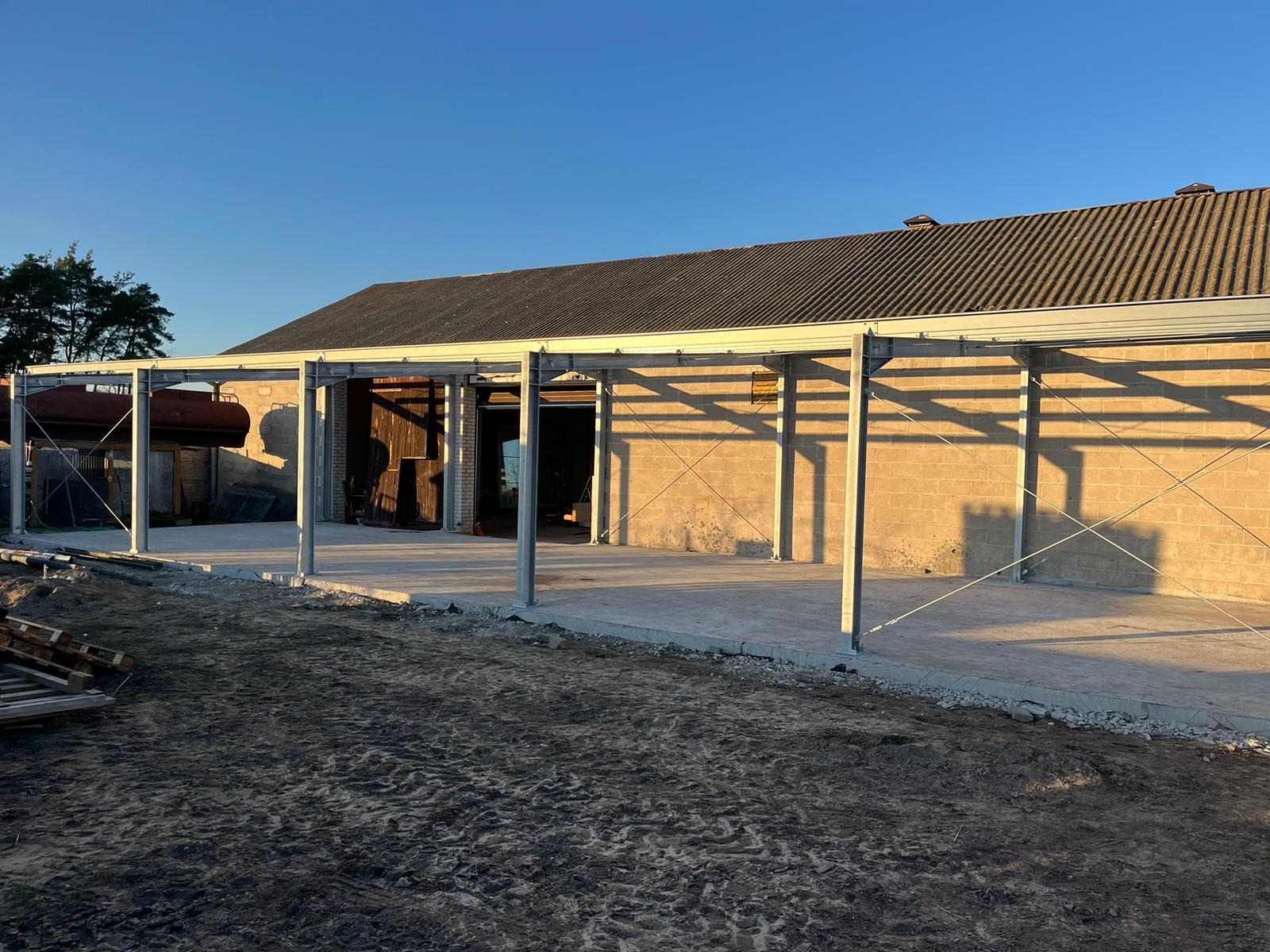Konstrukcja z metalowych słupów i belek na betonowej wylewce, prawdopodobnie zadaszenie lub wiata, w tle budynek z cegły i drewniane wrota, błękitne niebo.