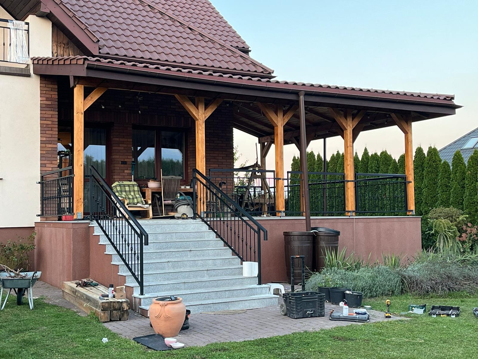 Zewnętrzne schody z szarego kamienia prowadzą na zadaszony taras z drewnianymi słupami i czarną, kutą balustradą. Widoczne narzędzia sugerują prace wykończeniowe.