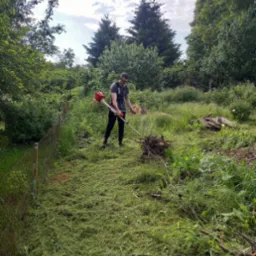 Mężczyzna w okularach, z podkaszarką spalinową, przycina trawę w ogrodzie, na tle drzew i nieba. Wokół leżą ścięte gałęzie.
