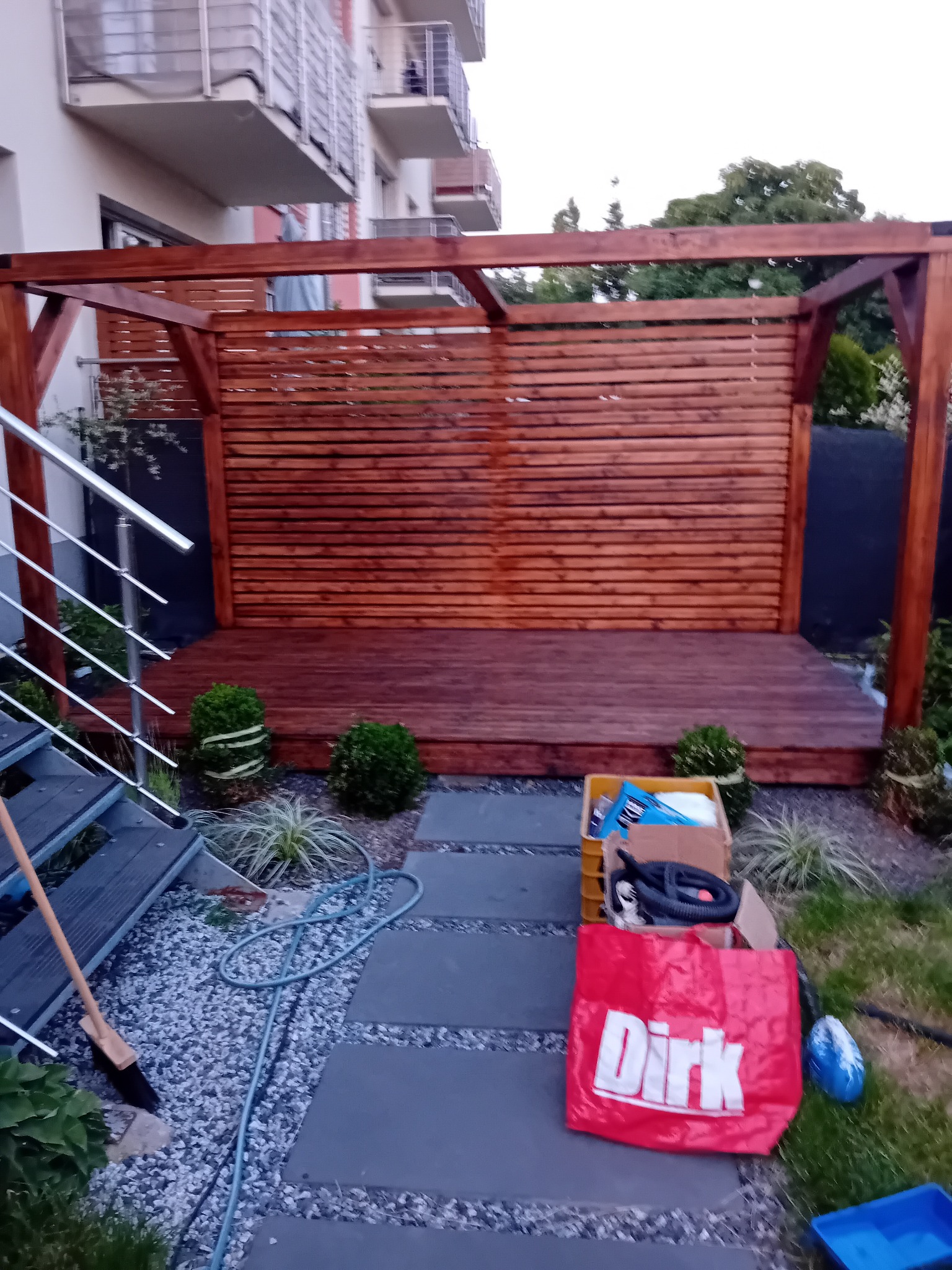 Zadaszona drewniana konstrukcja tarasowa z ażurową ścianką, widok z boku schodów. Obok narzędzia i akcesoria ogrodowe, kamienny chodnik i fragment trawnika.