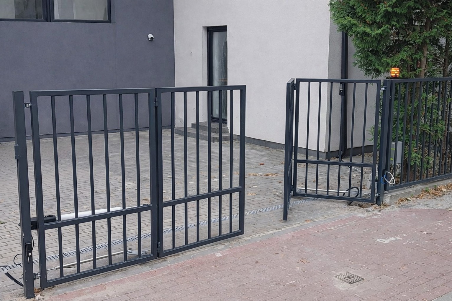 Otwarta, grafitowa brama wjazdowa z automatyką, widoczna na tle nowoczesnego domu z szarą elewacją i kostki brukowej. Brama z pionowymi szczeblami i poziomymi poprzeczkami.