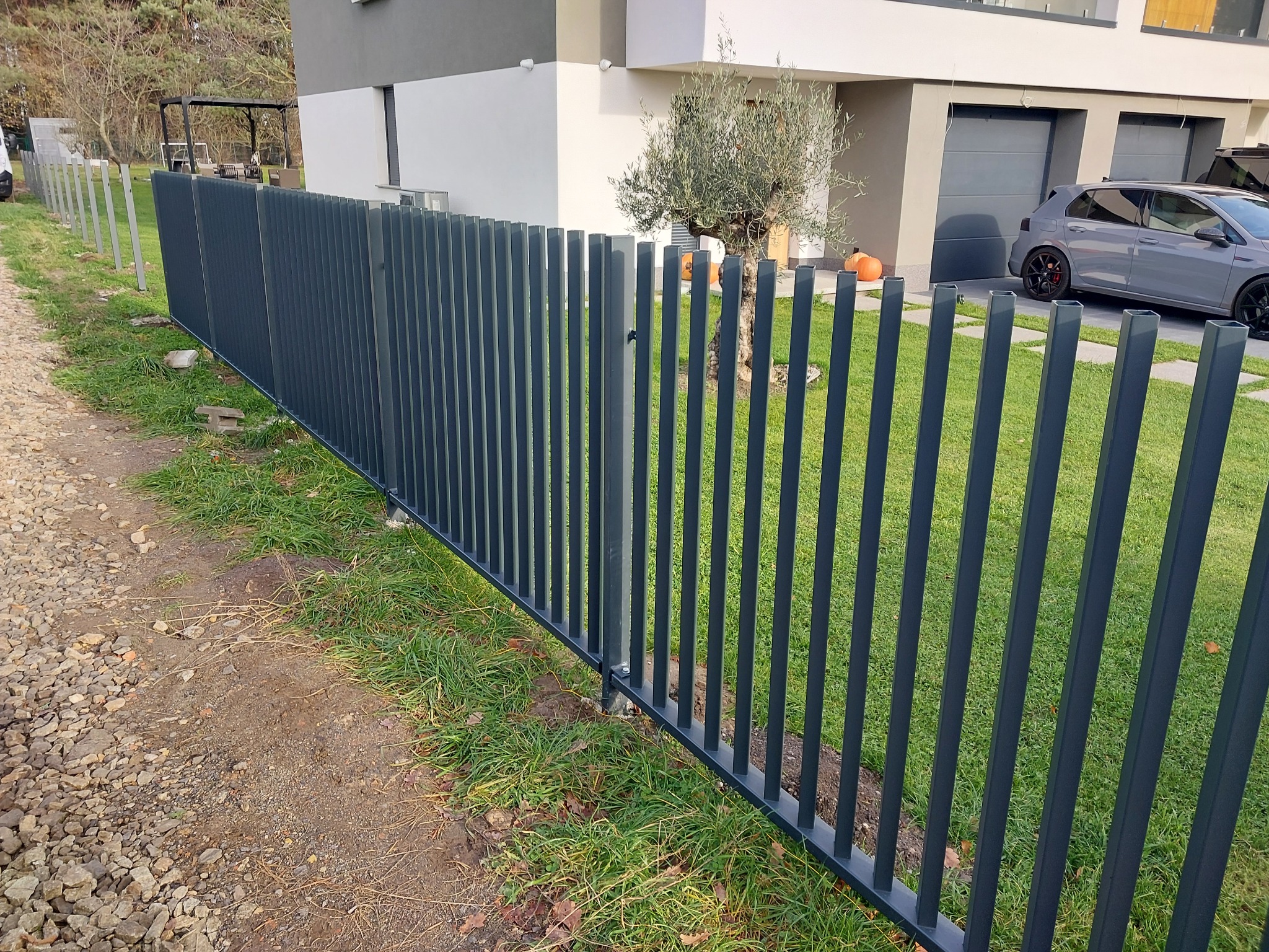 Nowoczesne, grafitowe ogrodzenie panelowe z kwadratowych profili, otaczające posesję z trawnikiem i domem z szarym autem w tle. Widok z poziomu gruntu.