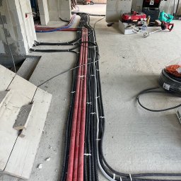 Daniel Krzyston - Kable elektryczne w peszlach ułożone równolegle na podłodze w surowym wnętrzu. Widoczne narzędzia i materiały budowlane. Instalacja w trakcie realizacji.