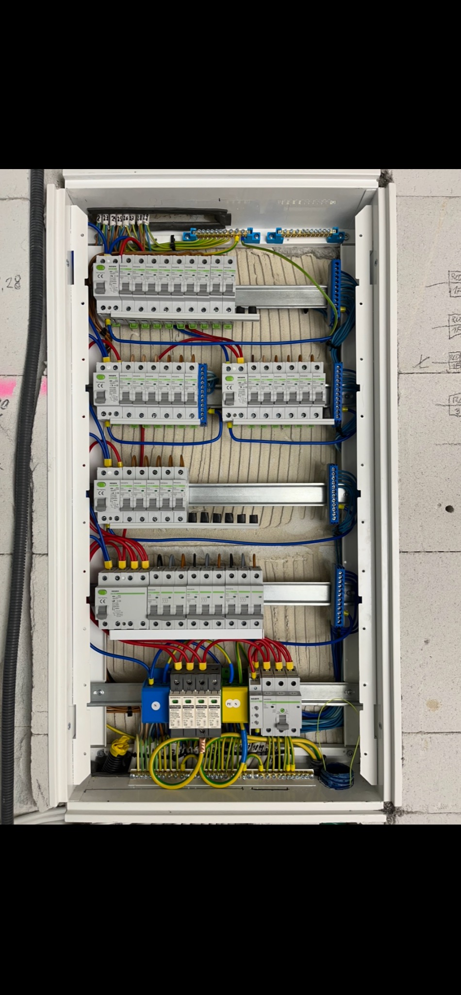 Rozdzielnia elektryczna w białej obudowie, widoczne bezpieczniki, przewody w kolorach: niebieski, czerwony, żółty, zielony. Instalacja zamontowana na ścianie z betonu komórkowego.