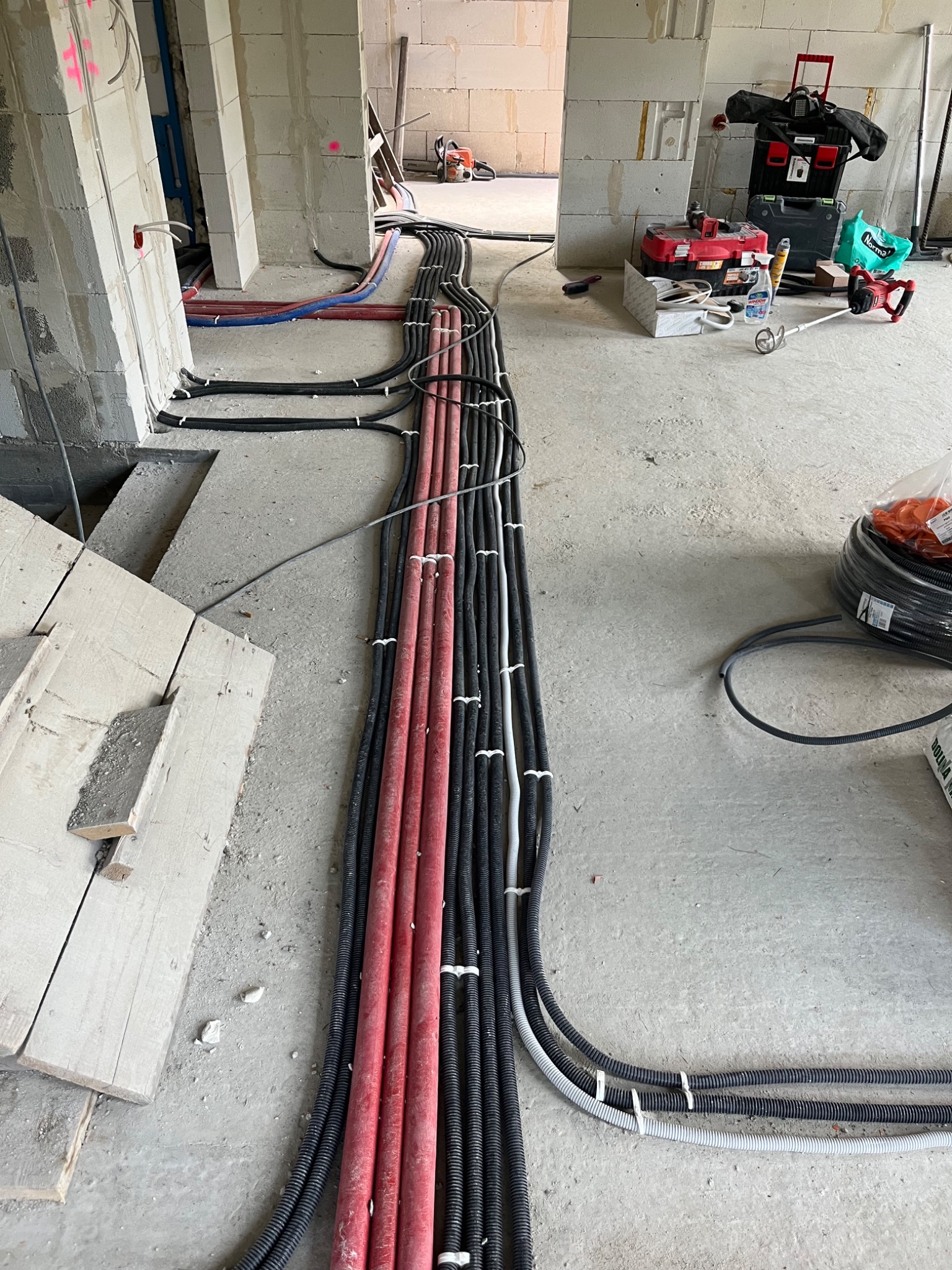Kable elektryczne w peszlach ułożone równolegle na podłodze w surowym wnętrzu. Widoczne narzędzia i materiały budowlane. Instalacja w trakcie realizacji.