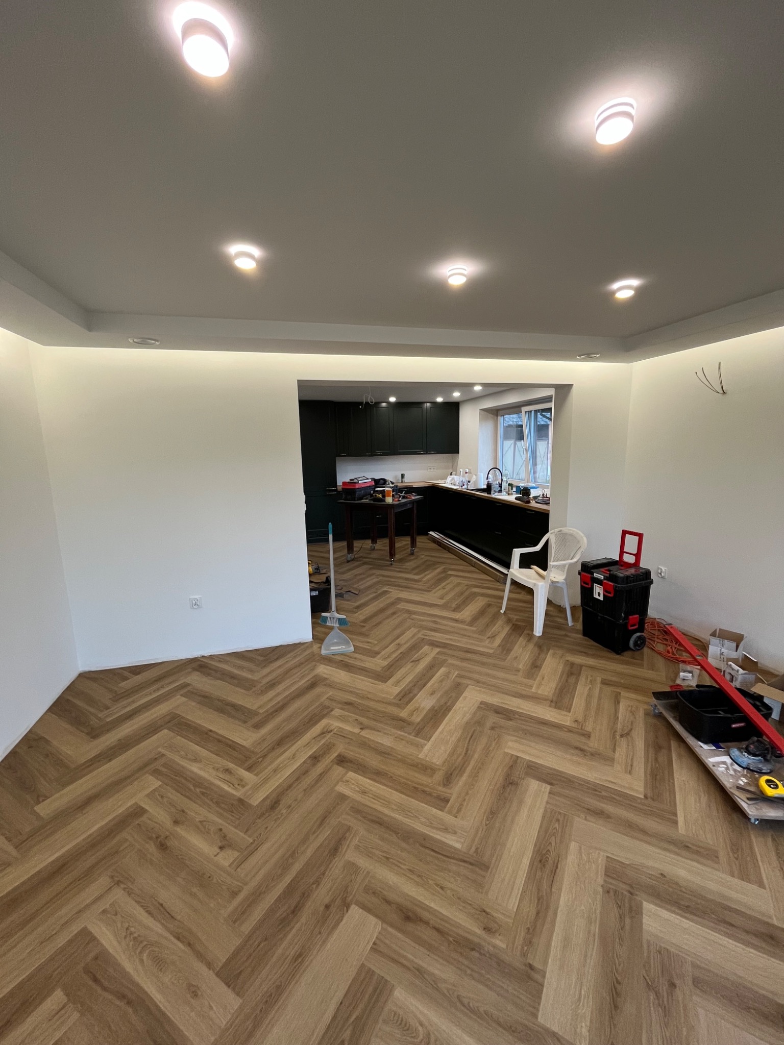 Wykończone wnętrze z parkietem w jodełkę, widok na kuchnię z czarnymi szafkami. Oświetlenie sufitowe punktowe i listwowe. Narzędzia pracy widoczne w tle.