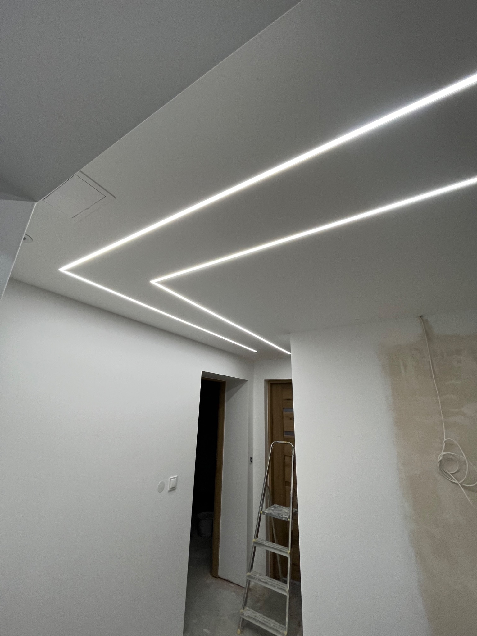 Białe wnętrze z podwieszanym sufitem i liniowym oświetleniem LED w kształcie zygzaka. Widoczna drabina i fragment ściany przygotowanej do malowania. Nowoczesny design.